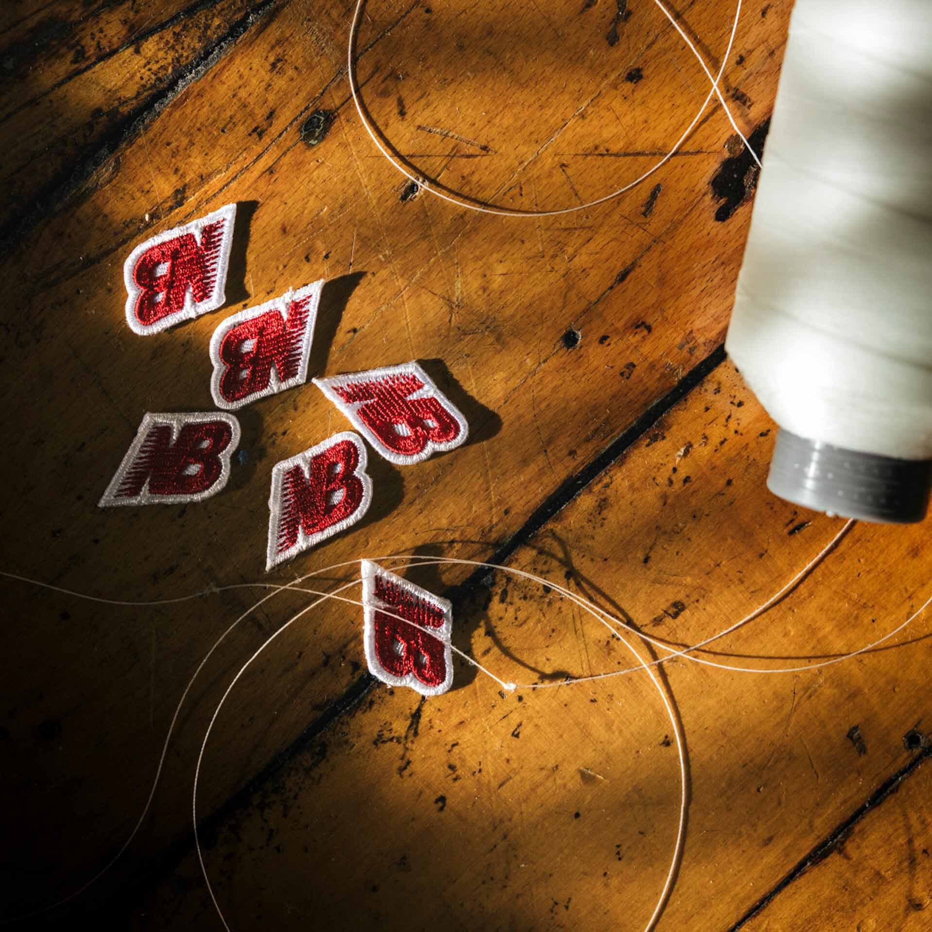 A spool of thread and multiple embroidered New Balance logos lying on a wooden table.