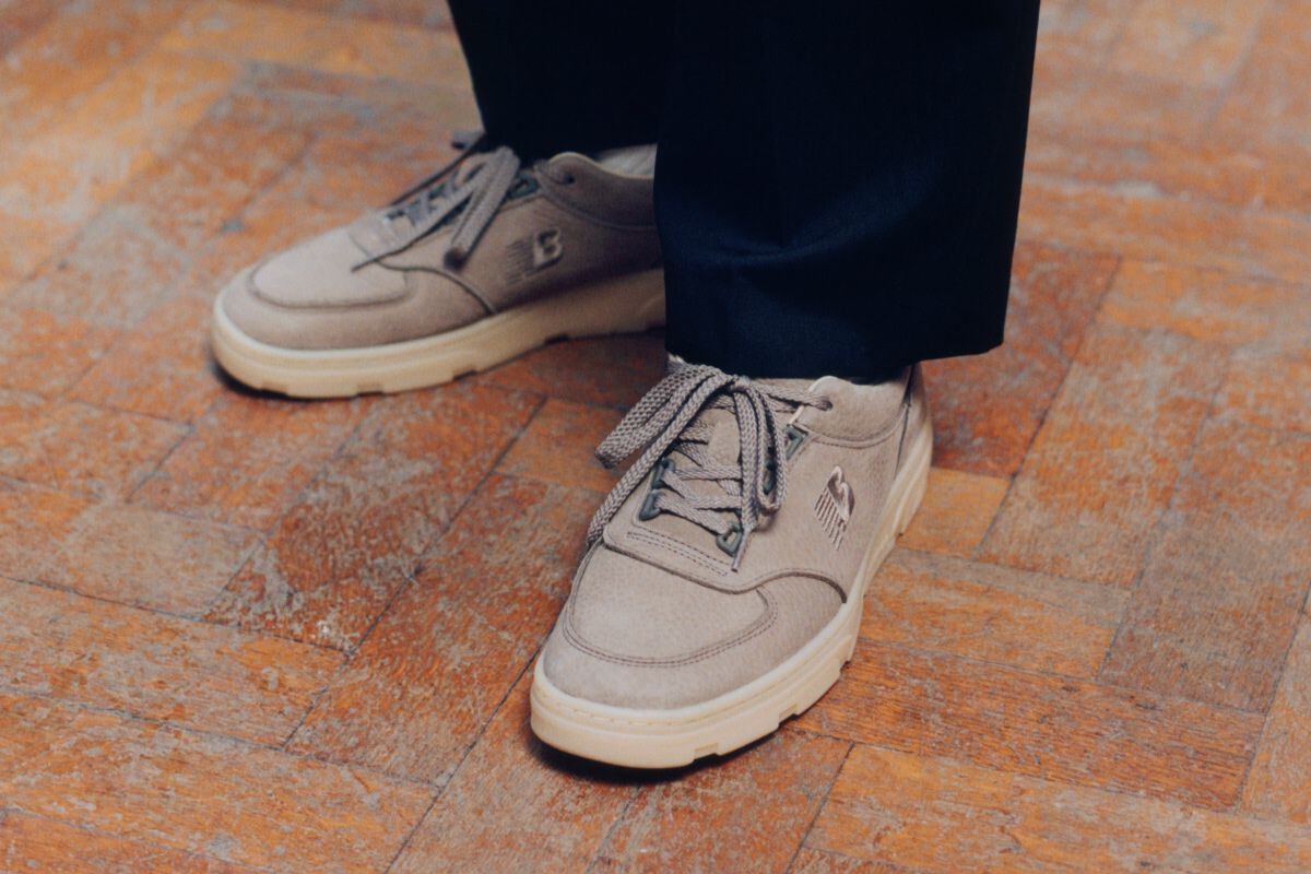 Knee down view of a man wearing a pair of New Balance Made in UK sneakers standing on a herringbone pattern floor with a white and pink rose being held in frame.
